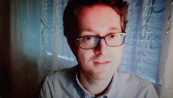 Ein junger Mann mit Brille und kurzen braunen Haaren lächelt leicht und blickt in die Kamera. Er trägt ein helles Hemd vor blauem Hintergrund.