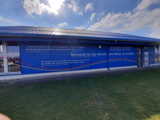 Blaues Gebäude mit Zitaten zur DDR-Grenzpolitik, Ulbrichts Mauerspruch und roten/gelben Linien unter sonnigem Himmel.