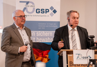 Zwei Männer präsentieren vor einem GSP-Banner zum 70-jährigen Jubiläum mit deutscher Flagge bei einer Veranstaltung.
