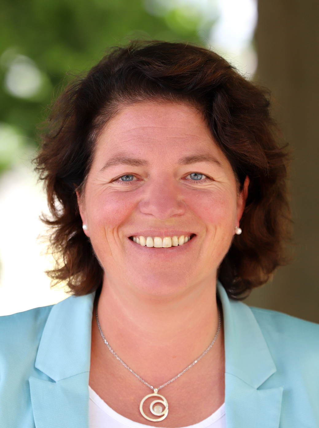Lächelnde Frau mit braunen Haaren, blauen Augen, hellblauem Blazer und Halskette vor grünem, unscharfem Hintergrund.