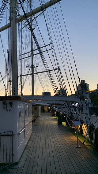 Blick vom Holzdeck eines Großseglers in der Dämmerung, mit Masten, Takelage und Stadtgebäuden vor warmem Himmel.