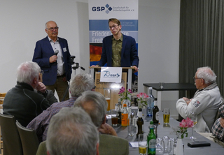 Zwei Männer sprechen auf einer GSP-Veranstaltung der Gesellschaft für Sicherheitspolitik vor einem Publikum älterer Männer an Tischen.