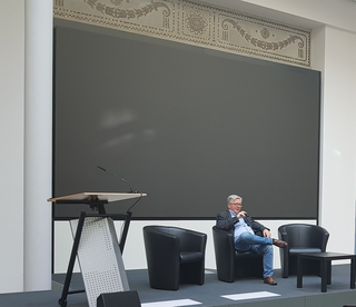 Ein Mann sitzt in einem Sessel auf einer Bühne mit Rednerpult und großem leeren Bildschirm, bereit für einen Vortrag oder Event.