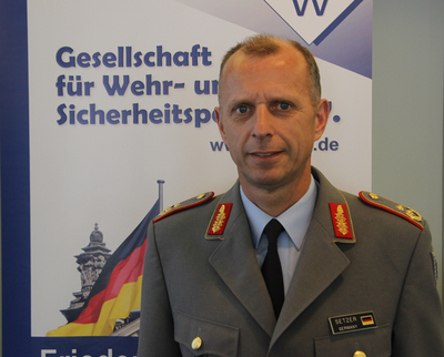 Mann in deutscher Militäruniform mit Namensschild 'SETZER, GERMANY' vor Banner der Gesellschaft für Wehr- und Sicherheitspolitik.