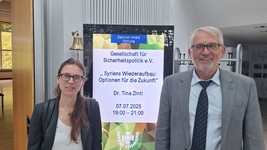 Zwei Personen vor einem Bildschirm, der einen Vortrag von Dr. Tina Zintl über Syriens Wiederaufbau ankündigt.