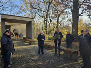 Männergruppe an einem Freilicht-Mahnmal mit Kränzen, einem Steinkreuz und Herbstbäumen. Ein Mann hält eine Topfpflanze.