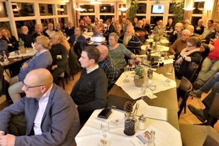 Eine Gruppe von Menschen sitzt an Tischen in einem hellen Restaurant oder Saal und nimmt an einer Veranstaltung teil.