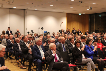 Großes professionelles Publikum, hauptsächlich Männer in Anzügen und einige Frauen, applaudiert bei einer Konferenz.