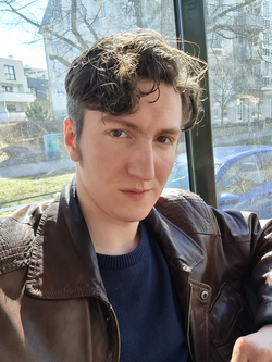 Eine junge Person mit welligem braunem Haar und Lederjacke blickt direkt in die Kamera, Sonnenlicht hebt Haare und Jacke hervor. Verwischter urbaner Hintergrund.