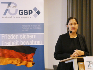 Frau spricht am Pult vor GSP-Banner mit Aufschrift 'Frieden sichern Freiheit bewahren' und deutscher Flagge.