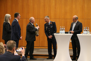 Ein älterer Mann übergibt ein Geschenk und spricht in ein Mikrofon zu einem Mann in Militäruniform bei einer Veranstaltung.