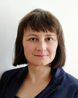 Eine Frau mit kurzen dunklen Haaren und einem blauen Blazer lächelt in die Kamera vor weißem Hintergrund.