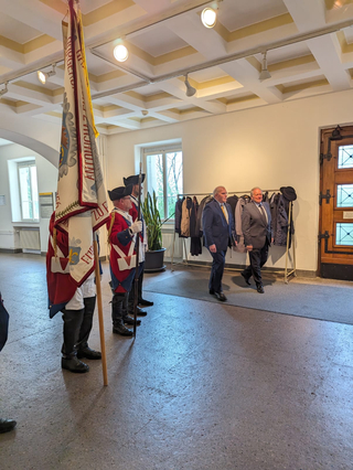 Zwei Männer in Anzügen gehen an drei Männern in historischen Uniformen vorbei, die eine große Fahne in einem Flur halten.