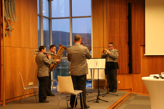 Ein militärisches Blechbläserquartett tritt drinnen auf, mit Posaune, Tuba und zwei Trompeten, vor einer Fensterfront.