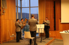 Ein militärisches Blechbläserquartett tritt drinnen auf, mit Posaune, Tuba und zwei Trompeten, vor einer Fensterfront.