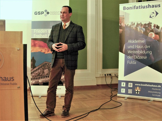 Mann hält Vortrag vor Bannern der Gesellschaft für Sicherheitspolitik und des Bonifatiushauses Fulda.