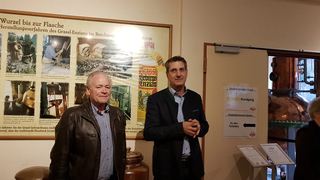 Zwei Männer in der Enzianbrennerei Grassl vor einem Info-Plakat zum Wurzel-bis-Flasche-Prozess und einem Brennkessel.