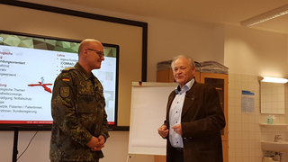 Ein Mann in Militäruniform und ein älterer Mann in Zivil unterhalten sich vor einem Präsentationsbildschirm.