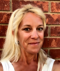 Eine lächelnde blonde Frau mit langen Haaren, dunklen Augen und einem weißen Oberteil vor einer roten Ziegelmauer.