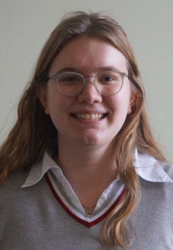 Lachende junge Frau mit Brille, langen braunen Haaren und grauem V-Pullover.