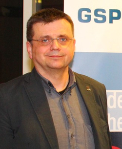 Lächelnder Mann mit Brille und dunkler Jacke vor einem Hintergrund mit 'GSP'-Schriftzug.