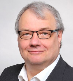 Porträt eines lächelnden Mannes mittleren Alters mit grauen Haaren, Brille, weißem Hemd und dunklem Sakko.