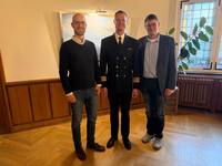 Drei lächelnde Männer, einer in Marineuniform, stehen in einem Raum mit Holzvertäfelung, Gemälde und Buntglasfenster.