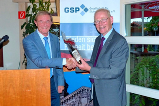 Zwei lächelnde Männer in Anzügen schütteln sich die Hände; einer überreicht eine Flasche als Geschenk bei GSP-Veranstaltung.