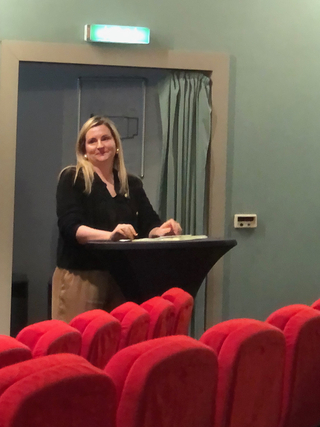 Lächelnde blonde Frau steht hinter einem schwarzen Rednerpult in einem Auditorium mit roten Sitzen.