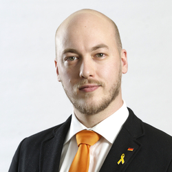 Kahlköpfiger Mann mit Bart, Anzug, orange Krawatte, Anstecknadeln der deutschen Flagge und einer gelben Schleife.