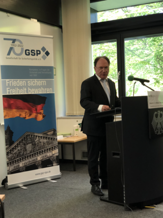 Mann im Anzug spricht an Rednerpult. GSP-Banner mit Reichstag, deutscher Flagge und Slogan 'Frieden sichern Freiheit bewahren' im Hintergrund.