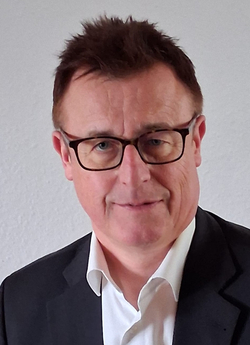 Porträt eines lächelnden Mannes mittleren Alters mit braunen Haaren, Brille, weißem Hemd und dunklem Sakko vor hellem Hintergrund.