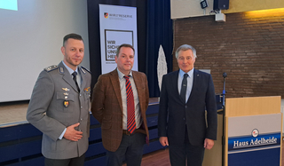 Drei Männer, darunter einer in Bundeswehr-Uniform, posieren vor einem Reservistenverband-Banner und einem Haus Adelheide Rednerpult.
