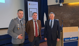 Drei Männer, darunter einer in Bundeswehr-Uniform, posieren vor einem Reservistenverband-Banner und einem Haus Adelheide Rednerpult.