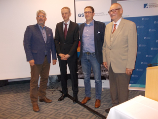 Vier Männer, teils in Anzügen, posieren vor Bannern der Konrad-Adenauer-Stiftung und der Gesellschaft für Sicherheitspolitik.