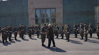 Militärmusikkapelle in Uniform, während zwei Männer, einer in Uniform, der andere in Zivil, sich die Hand geben.