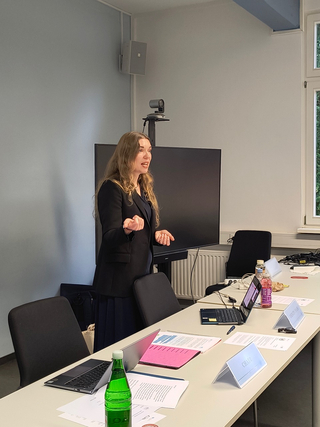 Eine Frau hält eine Präsentation, gestikulierend, in einem Konferenzraum mit Bildschirm, Laptops und Namensschildern auf einem Tisch.