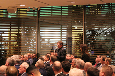 Mann spricht bei einer Konferenz ins Mikrofon, mit Publikum und großen Fenstern mit horizontalen Jalousien im Hintergrund.