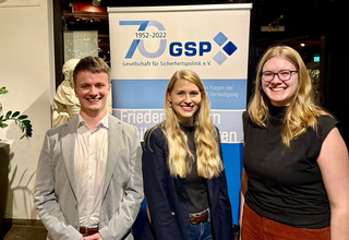 Drei junge Menschen, zwei Frauen und ein Mann, lächeln vor einem blauen GSP-Banner.