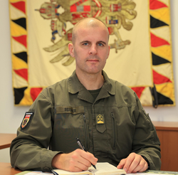 Kahlköpfiger Mann in olivgrüner österreichischer Militäruniform (Reisner), mit Stift und Notizbuch am Schreibtisch. Hintergrund: Flagge mit Adler.