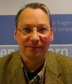 Mittelalter Mann mit Brille, Tweed-Sakko und kariertem Hemd vor blauem Hintergrund.