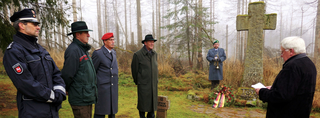 Gedenkzeremonie im nebligen Wald mit Offizieren, Polizist, Redner und Trompeter vor einem Weltkriegs-Kreuz.