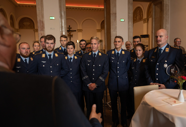 Eine Gruppe junger Militärangehöriger in dunkelblauen Uniformen steht aufmerksam einem Sprecher in einer geschmückten Halle gegenüber.