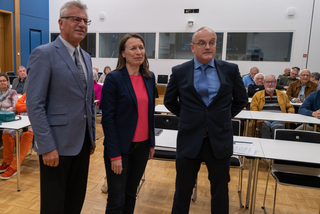 Drei Personen, zwei Männer und eine Frau, stehen vor einem Publikum an Tischen. Wahrscheinlich eine Präsentation.