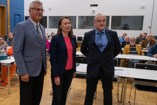 Drei Personen, zwei Männer und eine Frau, stehen vor einem Publikum an Tischen. Wahrscheinlich eine Präsentation.