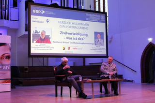Vortragsabend über Zivilverteidigung. Zwei Männer, einer in Zivil, einer in Uniform, sitzen vor einer Leinwand mit der Präsentation.