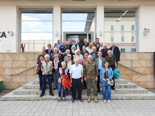 Gruppenfoto von Senioren auf einer Außentreppe vor einem Gebäude, darunter ein Mann in Militäruniform.
