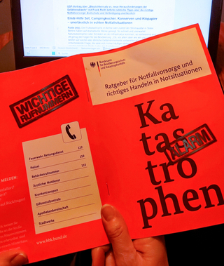 Eine Hand hält eine rote BBK-Broschüre 'Ratgeber für Notfallvorsorge' mit wichtigen Rufnummern und 'Katastrophen Alarm'.