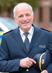 Ein lächelnder, grauhaariger Mann in deutscher Polizeiuniform, der seine Polizeimütze in der Hand hält.