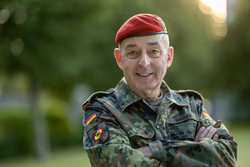 Ein lächelnder Soldat der Bundeswehr in Tarnuniform und rotem Barett, Arme verschränkt, vor grünem Hintergrund.